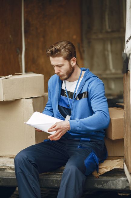 A man in a blue hoodie and dark trousers is sitting on the step of a moving truck or van, surrounded by cardboard boxes and packing materials. He is holding and reading from a sheet of paper, possibly a checklist or moving instructions, with a focused expression. The background shows a wooden interior environment, indicative of a property involved in home relocation. The scene captures the process of packing, organizing, or planning during a furniture transport and house removal. Various cardboard boxes, some sealed with tape, are stacked around him on the truck or van's loading area. The lighting appears natural and soft, highlighting the details of the boxes and the man's clothing. This visual illustrates the preparation stages of a house or office move, as undertaken by professional removals services, such as those offered by Man and Van Parsons Green, focusing on efficient packing and logistical planning for a successful relocation.