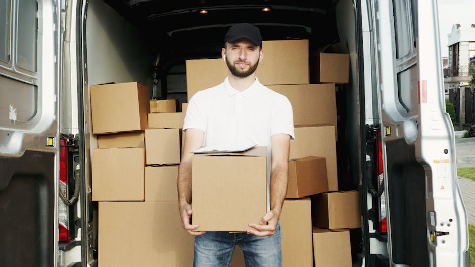A man wearing a black cap, white shirt, and jeans is standing inside the open rear doors of a white van, part of a house relocation process. He is holding a medium-sized cardboard box in front of him, with a clipboard resting on top of the box. The van's interior is filled with neatly stacked cardboard boxes of various sizes, some wrapped in plastic or packing materials, arranged to maximize space for efficient furniture transport and packing during the house move. Outside, there is a paved pathway and residential houses visible, indicating the vehicle is parked at a home or residential area, possibly on a pavement or driveway. Man and Van Parsons Green, specialising in removals, is involved in the loading process, which demonstrates effort and organisation in handling household items for an office or home relocation, consistent with professional moving services near Fulham Broadway.