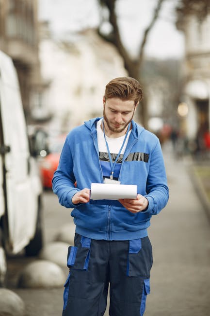 A man in a blue hoodie and dark trousers is sitting on the step of a moving truck or van, surrounded by cardboard boxes and packing materials. He is holding and reading from a sheet of paper, possibly a checklist or moving instructions, with a focused expression. The background shows a wooden interior environment, indicative of a property involved in home relocation. The scene captures the process of packing, organizing, or planning during a furniture transport and house removal. Various cardboard boxes, some sealed with tape, are stacked around him on the truck or van's loading area. The lighting appears natural and soft, highlighting the details of the boxes and the man's clothing. This visual illustrates the preparation stages of a house or office move, as undertaken by professional removals services, such as those offered by Man and Van Parsons Green, focusing on efficient packing and logistical planning for a successful relocation.