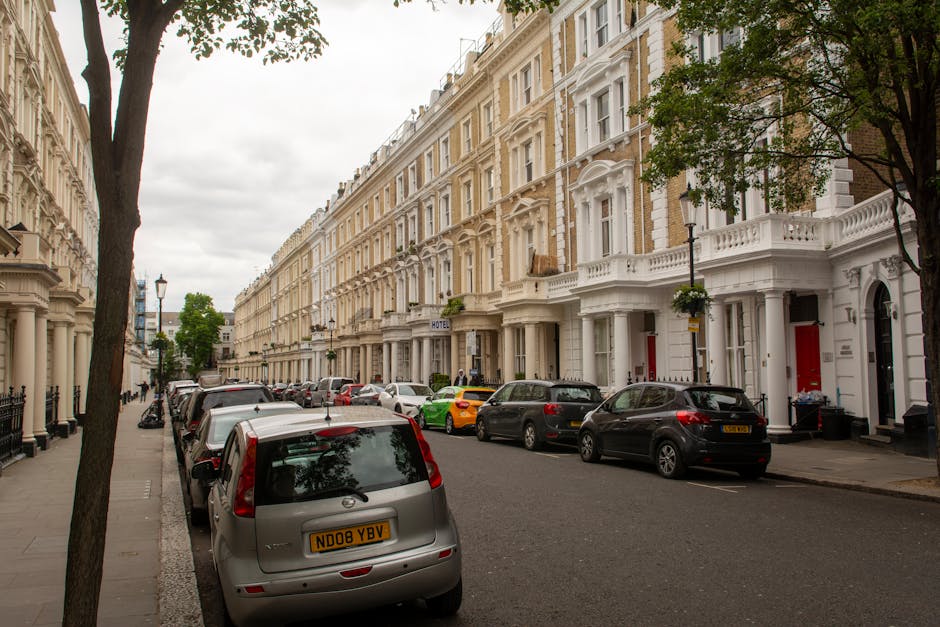 A row of Victorian terraced houses with ornate white facades and large windows lining a street in Parsons Green SW6, London, under an overcast sky. Several parked cars, including a silver van with the registration ND08 YBV, are positioned along the street. In front of one property, a man in casual clothing is maneuvering a trolley loaded with cardboard boxes and plastic-wrapped furniture, preparing for a home relocation. The scene captures the process of furniture transport and packing involved in a house removal, with the van positioned nearby for loading or unloading. Trees with green foliage partially obscure the upper stories of the buildings, which feature decorative balconies and white detailing. The area appears quiet, with minimal pedestrian activity, illustrating the typical environment for house removals in a residential London neighbourhood, as handled by Man and Van Parsons Green.