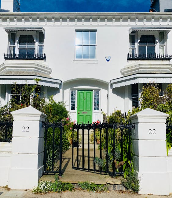 A two-storey white Victorian terraced house located at 22 Parsons Green SW6, featuring a vibrant green front door centered between two large bay windows with decorative black wrought iron railings. The building has a detailed cornice along the roofline and symmetrical balcony railings on the upper floor. In front of the house is a low white brick wall with two matching pillars, each displaying the house number '22,' and a black metal gate opening onto a narrow paved pathway leading to the entrance. The pathway is flanked by well-maintained garden beds with various green plants and flowering shrubs, indicating recent gardening work. The scene is captured in natural daylight, highlighting the neat, classic architectural details of the property, which may be part of a home relocation or furniture transport process supervised by Man and Van Parsons Green, as part of their house removals services.