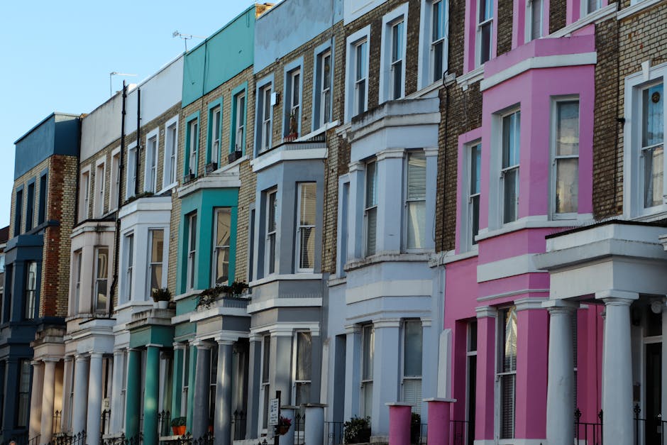 A row of Victorian terraced houses with ornate white facades and large windows lining a street in Parsons Green SW6, London, under an overcast sky. Several parked cars, including a silver van with the registration ND08 YBV, are positioned along the street. In front of one property, a man in casual clothing is maneuvering a trolley loaded with cardboard boxes and plastic-wrapped furniture, preparing for a home relocation. The scene captures the process of furniture transport and packing involved in a house removal, with the van positioned nearby for loading or unloading. Trees with green foliage partially obscure the upper stories of the buildings, which feature decorative balconies and white detailing. The area appears quiet, with minimal pedestrian activity, illustrating the typical environment for house removals in a residential London neighbourhood, as handled by Man and Van Parsons Green.