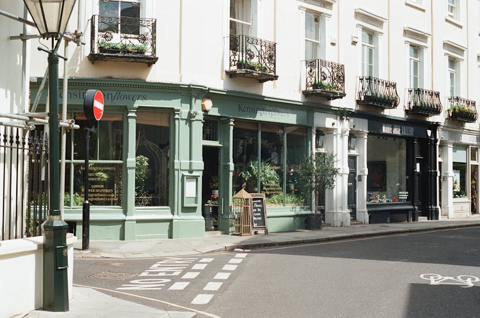 A street scene outside a shop located in a multi-storey building with white facade walls and black wrought iron balcony railings on the upper floors. The shop has large glass windows displaying potted