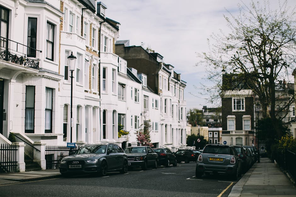 Image showing a residential street with white multi-storey houses featuring bay windows, decorative detailing, and small front gardens with black iron railings. Several parked cars, including an Audi and a Mini, line the street alongside the curb, which is partly composed of a grey pavement. A tall, black street lamp is positioned near the kerb, and a large tree with budding leaves extends over the sidewalk, indicating a springtime setting. The sky is overcast, providing diffuse lighting to the scene. This environment depicts an urban area suitable for home relocation or furniture transport activities, with the vehicles and property layout illustrating typical street parking conditions for house removals. Occasionally, moving equipment such as trolleys or packing materials may be used in such settings by professional removals services like Man and Van Parsons Green, to facilitate a smooth loading process from the property to the vehicle, which is visible parked nearby.