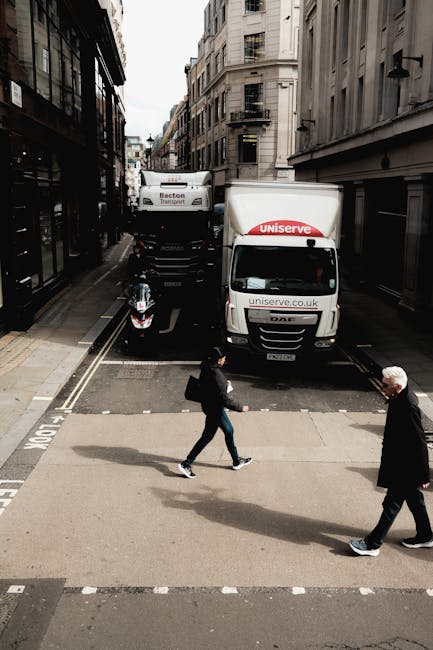 An urban street scene showing a narrow road with tall, multi-storey buildings on both sides. Two Man and Van Parsons Green vehicles are parked parallel to the kerb, with one white van displaying the 'uniserve' logo and website, indicating professional furniture transport and home relocation services. A motorcycle is parked beside the vans, close to the pedestrian crossing. In the foreground, two pedestrians—one man with white hair and dark clothing, and a woman in casual attire carrying a black bag—are crossing the street at a pedestrian crossing marked with white dashed lines. Overcast natural lighting highlights the building facades and street activity, illustrating a typical setting for packing, loading, and moving logistics involved in house removals, with the vehicles positioned for loading or unloading furniture and packing materials.