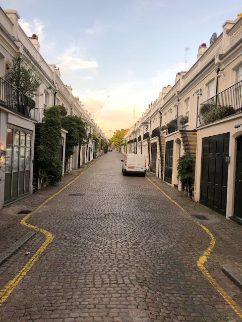 A narrow cobblestone street in Parsons Green with white terraced houses on both sides, each featuring small front gardens with green shrubs and potted plants. A white van is stationary near the center of the street, with its back door open, indicating a loading or unloading process. The street is lined with yellow markings, with a few outdoor tables and doors visible at ground level. The sky above is partly cloudy with soft, natural light illuminating the scene, reflecting typical residential surroundings suited for house removals and furniture transport facilitated by Man and Van Parsons Green during a home relocation.