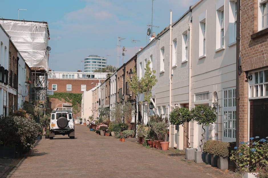 A narrow cobblestone street in Parsons Green with white terraced houses on both sides, each featuring small front gardens with green shrubs and potted plants. A white van is stationary near the center of the street, with its back door open, indicating a loading or unloading process. The street is lined with yellow markings, with a few outdoor tables and doors visible at ground level. The sky above is partly cloudy with soft, natural light illuminating the scene, reflecting typical residential surroundings suited for house removals and furniture transport facilitated by Man and Van Parsons Green during a home relocation.
