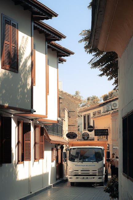 A white moving truck parked on a narrow street between residential buildings with light-colored facades and brown wooden shutters. The truck is positioned near the entrance of a property, with part of the vehicle inside a courtyard or driveway. Bright sunlight casts shadows on the buildings, and some foliage from nearby trees partially extends into the frame. In the background, a person is visible near the truck, likely involved in loading or unloading furniture and boxes. The scene depicts an ongoing home relocation or furniture transport operation facilitated by a professional removals service, with visible packing materials like cardboard boxes and blankets perhaps on or near the truck, supporting a smooth loading process. The setting aligns with typical conditions for house removals in an urban or suburban area, emphasizing the importance of careful handling of belongings during packing and moving procedures. The image visibly captures the logistics and environment associated with furniture transport, as part of the broader moving services offered by Man and Van Parsons Green as detailed on their website.