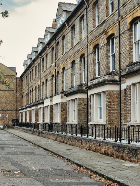 A row of traditional and older brick terrace houses with multiple floors, each featuring large sash windows with white frames and small balconies or window ledges; the buildings are separated from a narrow sidewalk by short black metal railings, with some windows partially covered by protective plastic or curtains. The street is paved with asphalt, with a slight incline and some fallen leaves scattered along the pavement. The sky is overcast, casting diffused light over the scene. This exterior setting illustrates an urban residential area suitable for home relocation or furniture transport, relevant to professional removals services like those offered by Man and Van Parsons Green, with an emphasis on efficient loading or unloading processes outside similar properties on New Kings Road.