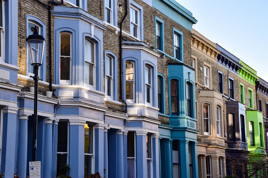 A row of traditional and older brick terrace houses with multiple floors, each featuring large sash windows with white frames and small balconies or window ledges; the buildings are separated from a narrow sidewalk by short black metal railings, with some windows partially covered by protective plastic or curtains. The street is paved with asphalt, with a slight incline and some fallen leaves scattered along the pavement. The sky is overcast, casting diffused light over the scene. This exterior setting illustrates an urban residential area suitable for home relocation or furniture transport, relevant to professional removals services like those offered by Man and Van Parsons Green, with an emphasis on efficient loading or unloading processes outside similar properties on New Kings Road.