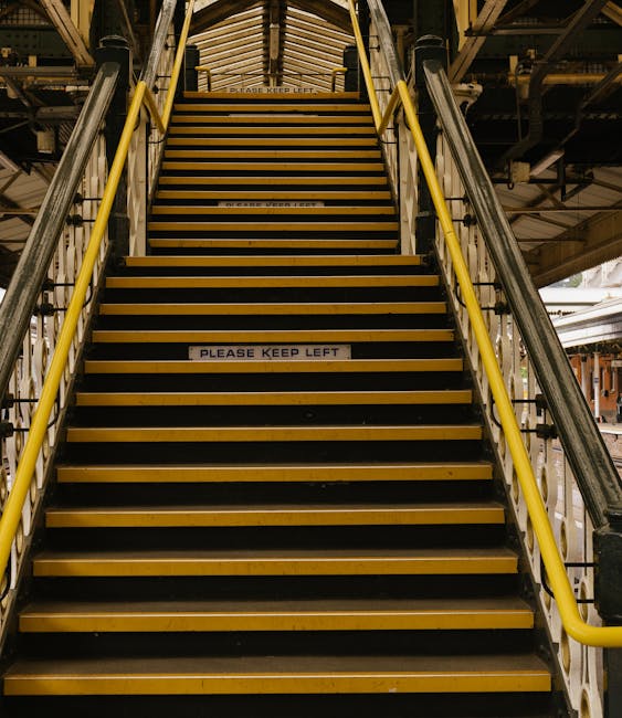 A set of indoor metal stairs with yellow safety handrails on each side, leading to an upper level in a building that appears to be part of a train station or industrial facility, with visible structural beams and lighting fixtures overhead. The stairs are covered with non-slip black strips along the treads, and small signs on some steps read 'Please Keep Left'. The environment is well-lit, and at the bottom of the stairs, there is a paved floor area extending into the background. This image relates to house removals and moving logistics, as it depicts the kind of access points that might be involved in furniture transport and home relocation processes, supported by the services of Man and Van Parsons Green.