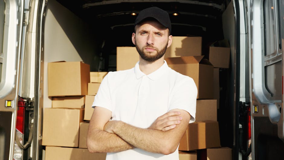 A man standing inside the open rear doors of a van loaded with multiple cardboard boxes of various sizes, arranged on the floor and against the walls of the vehicle. The man has a beard, is wearing a black cap, a white short-sleeved shirt, and is crossing his arms while looking directly at the camera with a neutral expression. The scene is set in daylight, with the van parked on the pavement, and the background showing a clean, neutral environment typical of house removals. The cardboard boxes are sealed with packing tape, indicating they are prepared for moving or home relocation. The interior of the van is dark, contrasting with the exterior lighting, and the focus is on the man and the packed boxes, reflecting the process of furniture transport, packing, and loading involved in professional moving services like those offered by Man and Van Parsons Green.