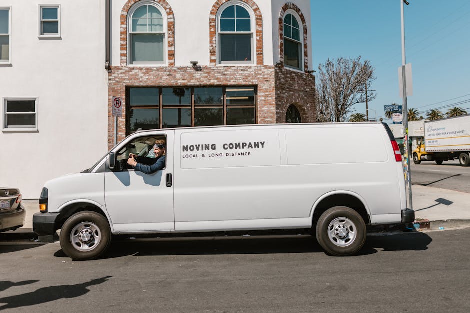 A white moving van with the text 'Moving Company Local & Long Distance' written on its side is parked on a street in front of a multi-storey brick building with large arched windows. The van's driver, wearing a dark jacket and sunglasses, is sitting in the vehicle, holding the steering wheel, and appears to be preparing for a home relocation or furniture transport. Behind the van, a few cardboard boxes wrapped in plastic or fabric are visible in the back, possibly packed for a house move. The surroundings include a few parked cars, a tree, and other vehicles, with clear weather and daylight illuminating the scene. This image captures the loading process typical of professional removals services, such as those provided by Man and Van Parsons Green, indicating a quick and organized move procedure, consistent with same-day moving operations in Parsons Green.