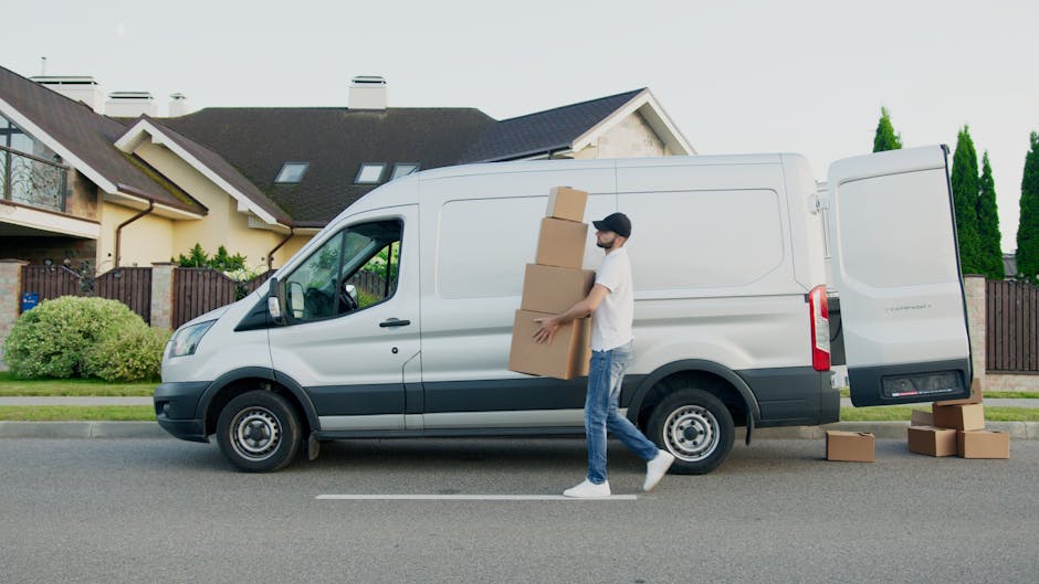 A man standing inside the open rear doors of a van loaded with multiple cardboard boxes of various sizes, arranged on the floor and against the walls of the vehicle. The man has a beard, is wearing a black cap, a white short-sleeved shirt, and is crossing his arms while looking directly at the camera with a neutral expression. The scene is set in daylight, with the van parked on the pavement, and the background showing a clean, neutral environment typical of house removals. The cardboard boxes are sealed with packing tape, indicating they are prepared for moving or home relocation. The interior of the van is dark, contrasting with the exterior lighting, and the focus is on the man and the packed boxes, reflecting the process of furniture transport, packing, and loading involved in professional moving services like those offered by Man and Van Parsons Green.