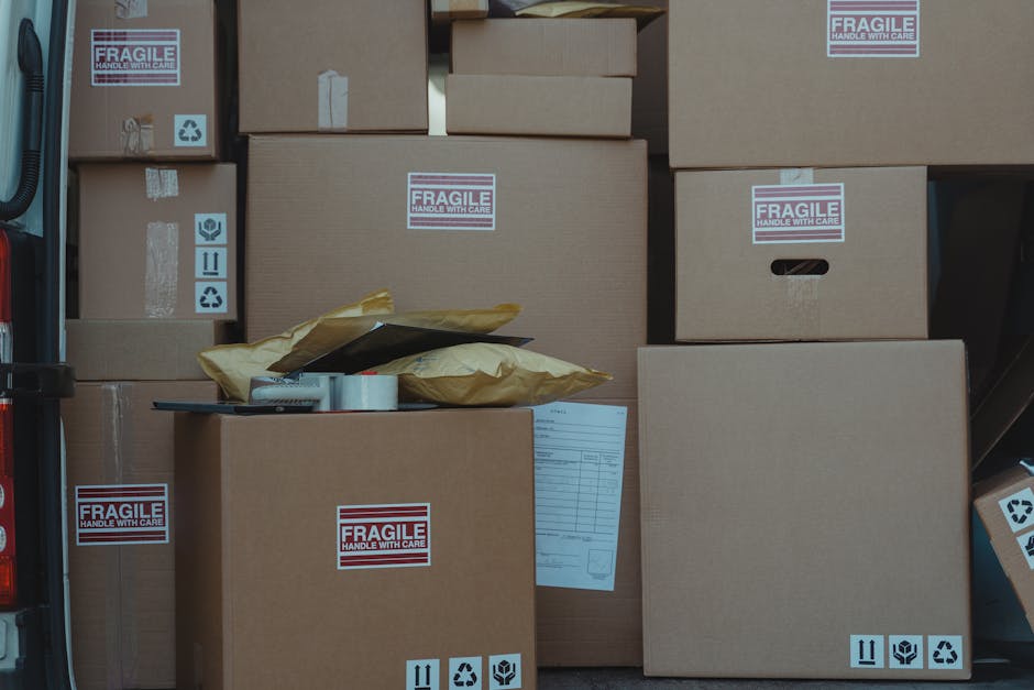 A collection of various cardboard boxes stacked inside a vehicle or on a loading area, with several labeled 'FRAGILE HANDLE WITH CARE.' Some boxes are sealed with packing tape, and others have printed
